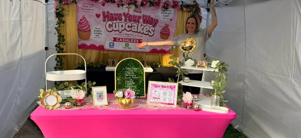 The Have Your Way Cupcakes booth inside a white tent: a pink table with flowers, a flavor board, a pay-by-QR sign, and a pink banner with the Have Your Way Cupcakes logo, with the owner smiling behind the table.